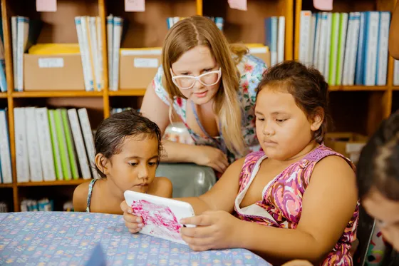 A facilitator and girls reading a tablet in front of library shelves