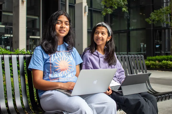 Two students on a bench, each with her own device
