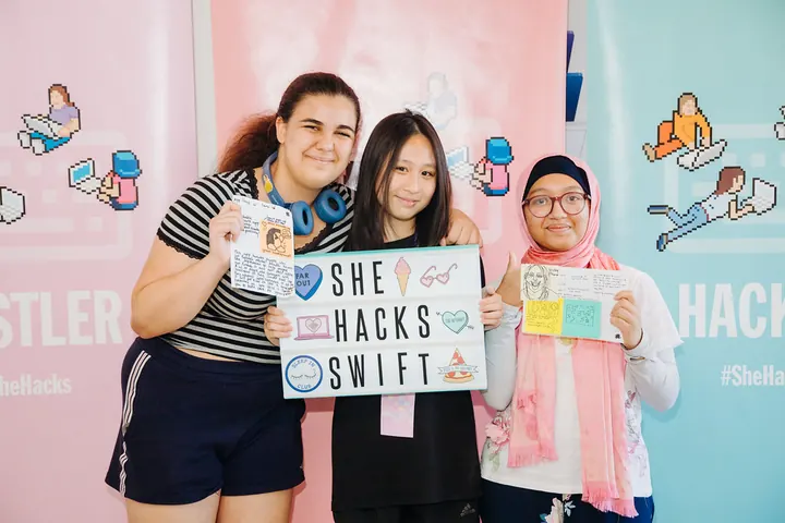 Three students holding up signs, one reading SheHacksSwift